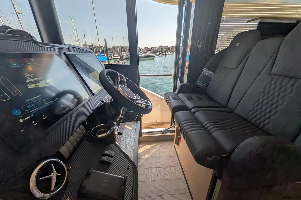 Sleek motorboat cockpit featuring digital controls, steering wheel, and plush seating, overlooking a marina with boats and water.